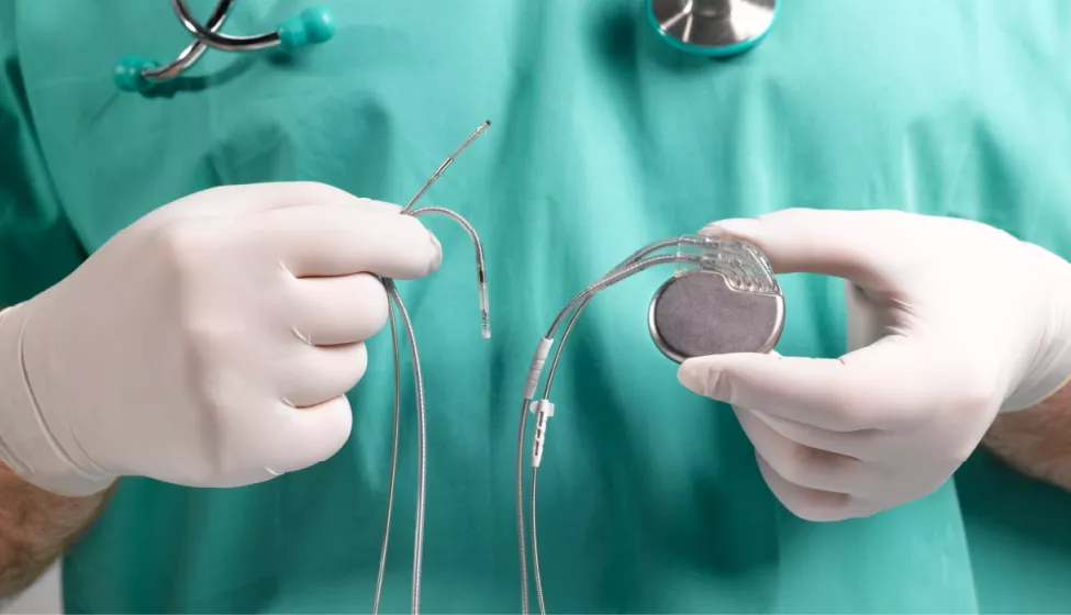 Gloved, medical professional in green scrubs with stethoscope around neck (shown from neck to mid torso) holding an implanted medical device